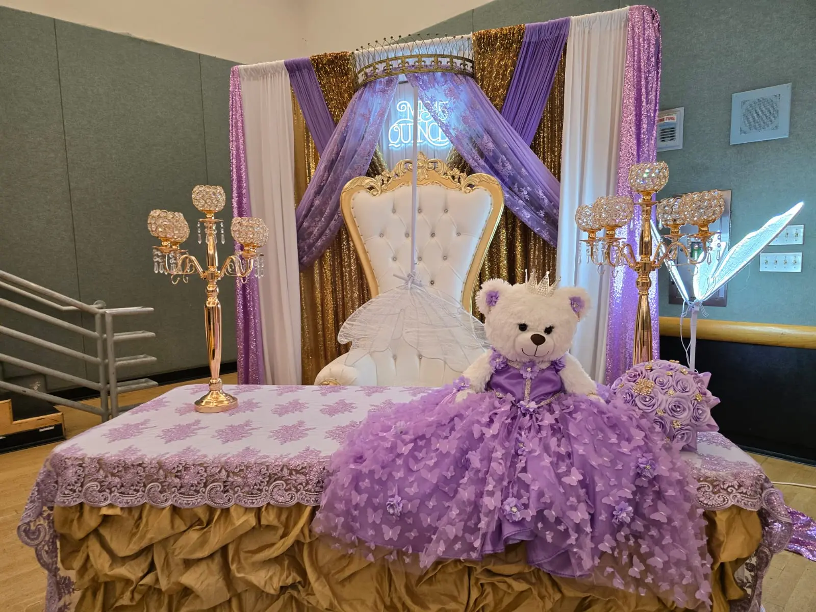Purple quinceañera setup with gold draping, crown canopy, throne chair and lace table