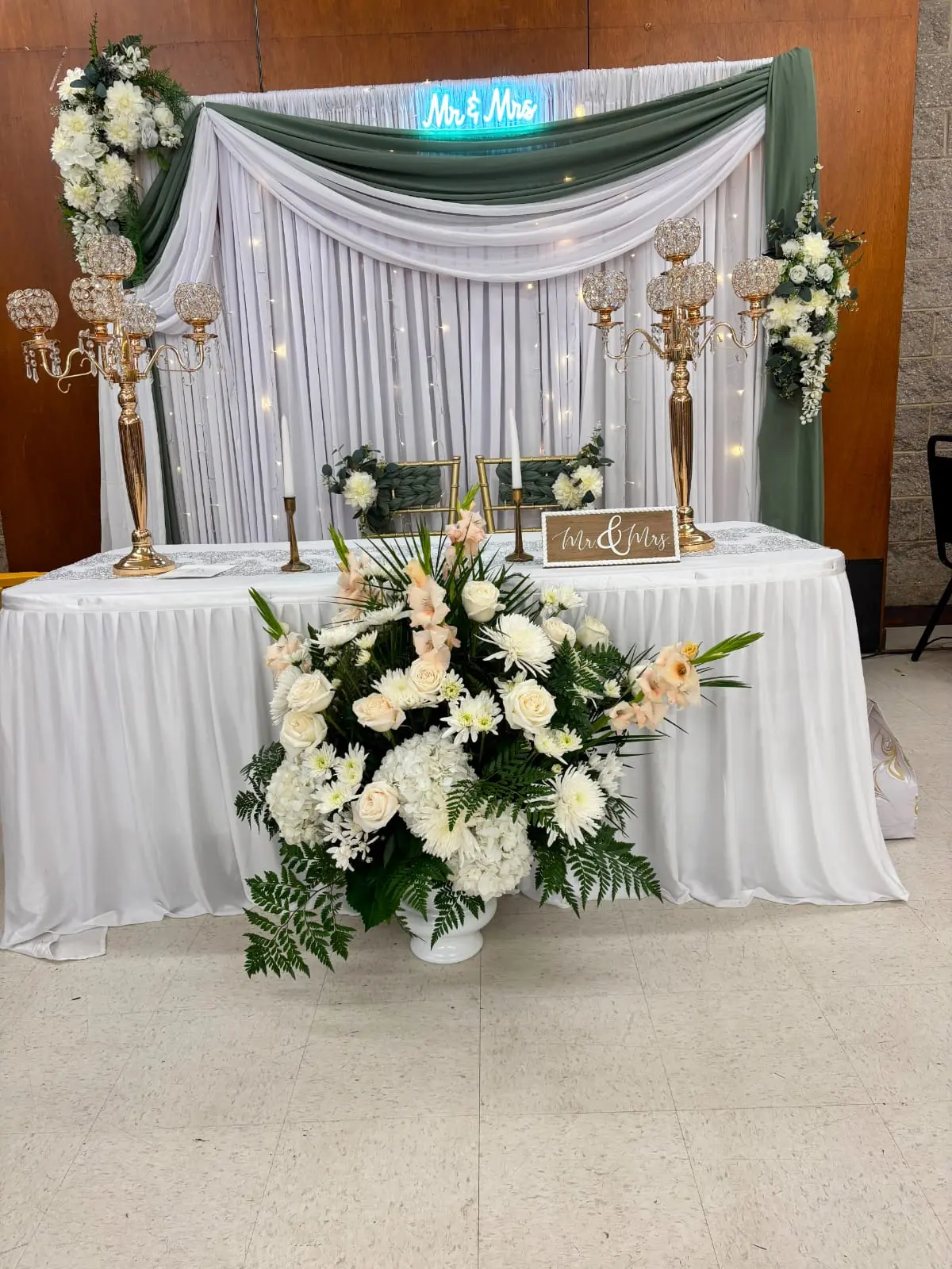 White and sage green draped head table with candelabras, white florals and a Mr & Mrs neon sign