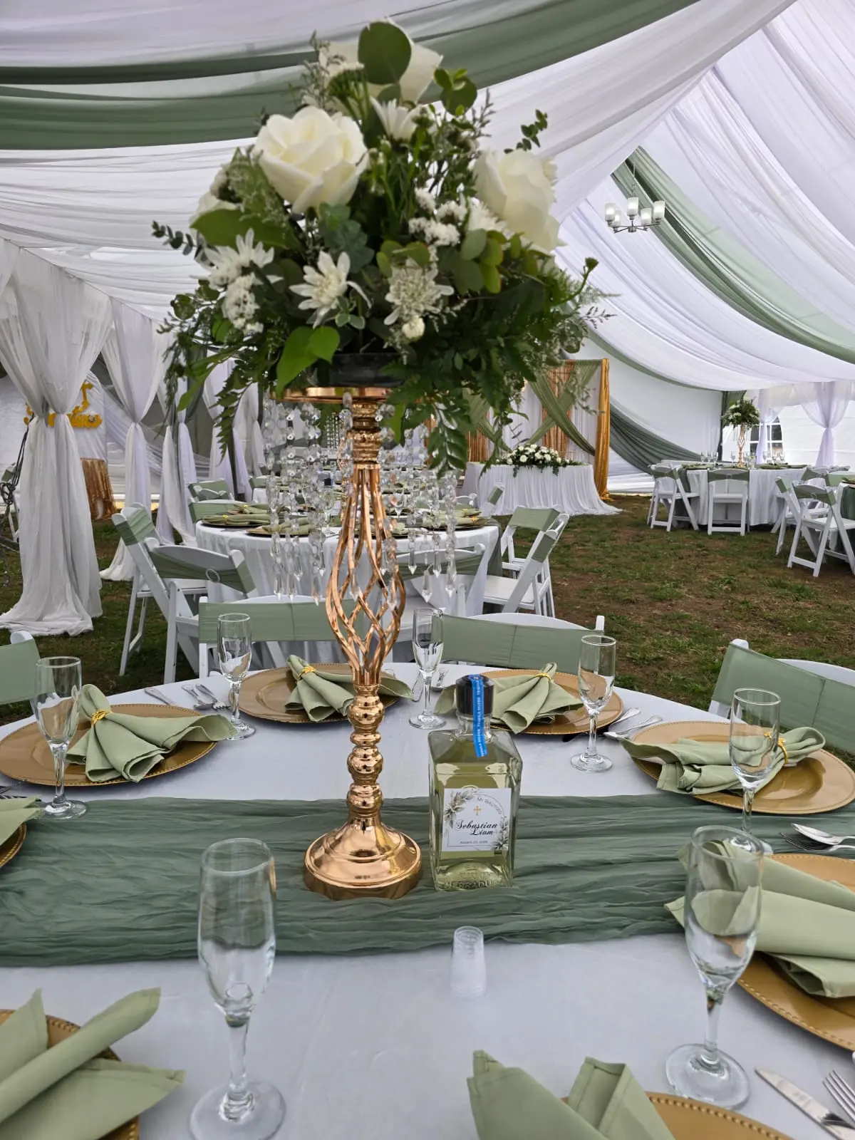 Tent with white and sage draping, tall floral centerpiece and gold chargers with sage napkins